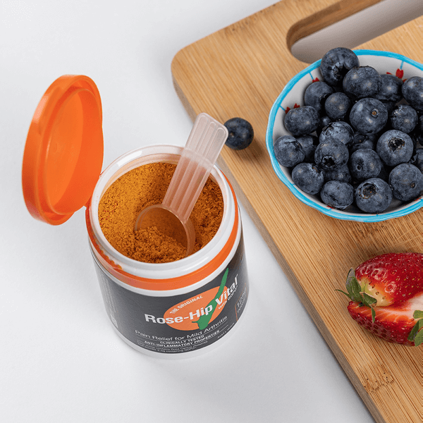 Open jar of Rose-Hip Vital with a scoop and bowl of blueberries on a wooden cutting board.